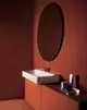 Weißes Waschbecken auf einer oranger Holzablage, beleuchtet von reflektierendem Licht, mit runder Wandspiegel und minimalistischen Pflegeprodukten, entworfen von Ideal Standard.